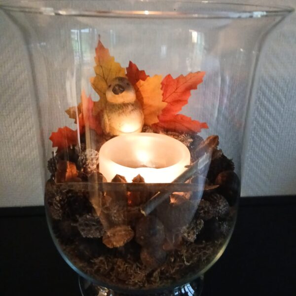 Photophore sur pied en verre transparent, décoré de mousse, de pommes de pin, de bois et de feuilles d’automne, avec une figurine d’oiseau.
