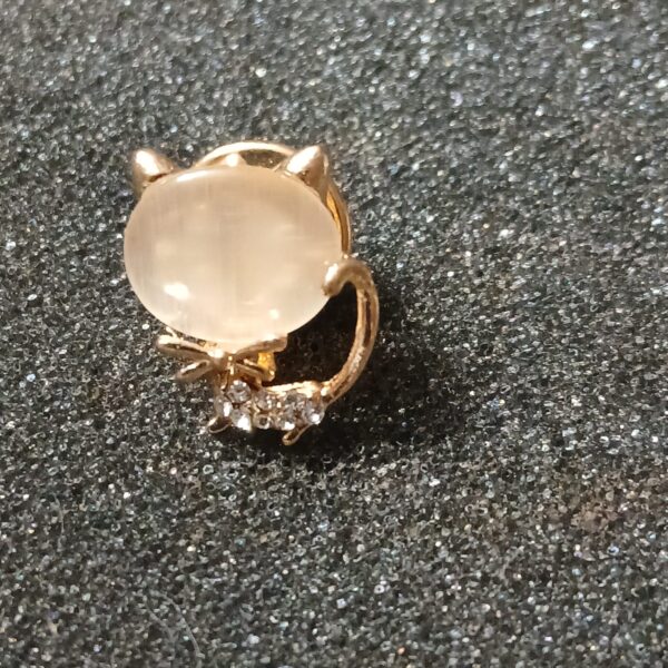 Petite broche représentant un chat stylisé au corps d’aspect nacré, avec des détails dorés ornés de petits strass.